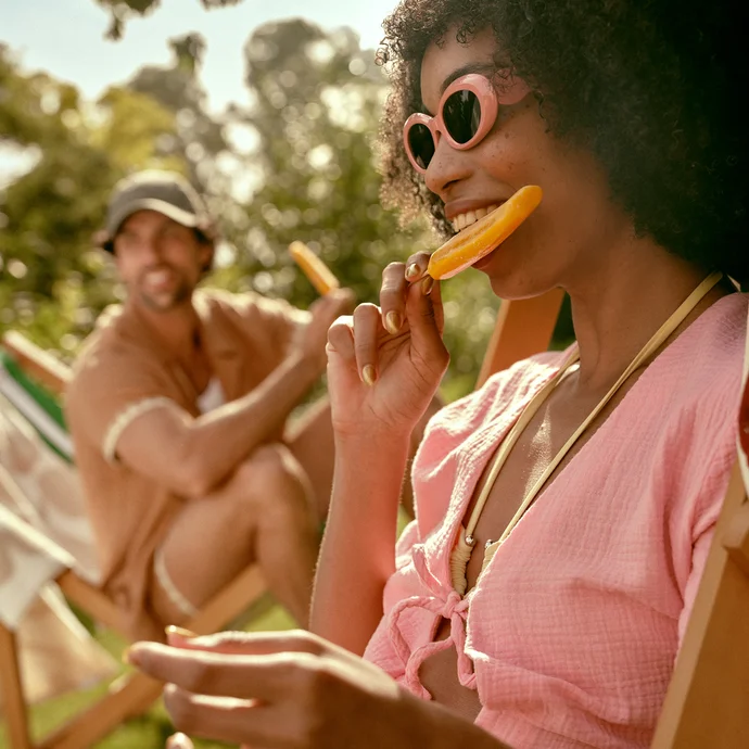 A woman wearing sunglasses smiling and eating an orange popsicle outdoors.
