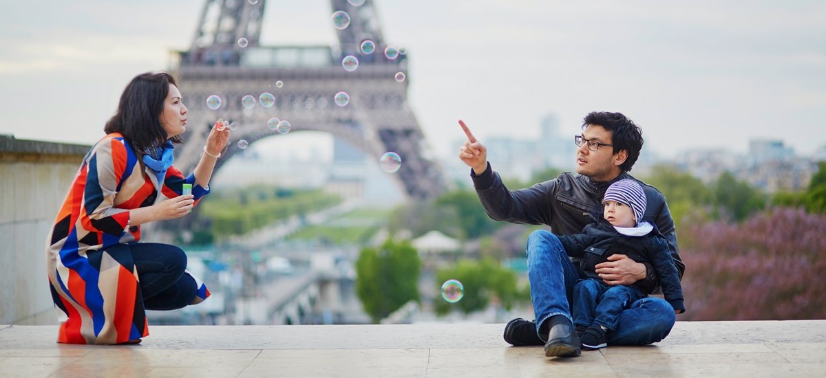 Young family sitting together, mother blowing bubbles while father and child look on. Eiffel tower in background