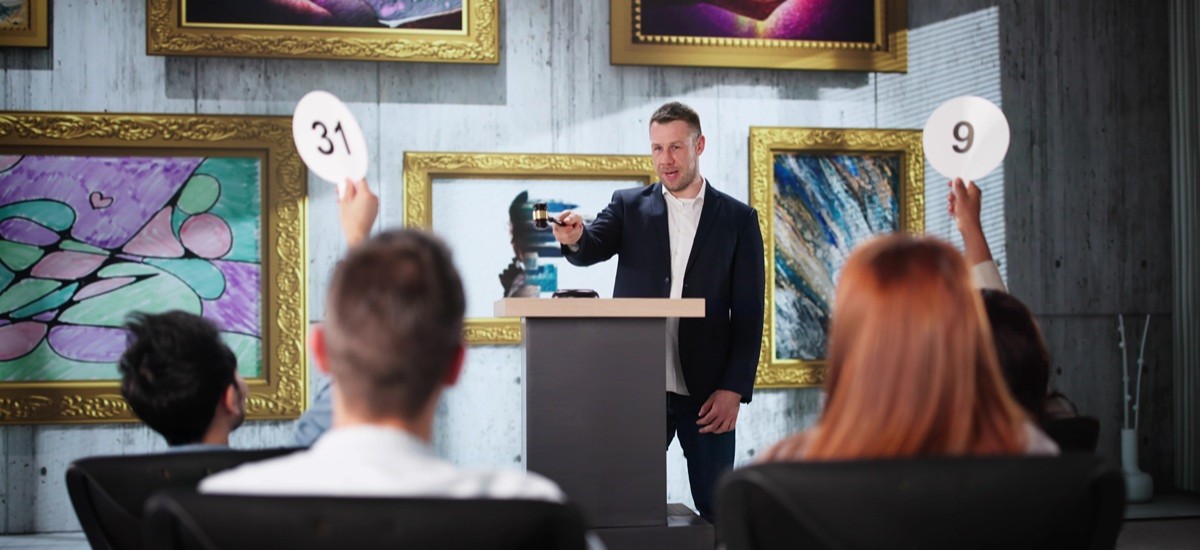 An auctioneer takes bids for art in a auction house, several people have their paddles raised
