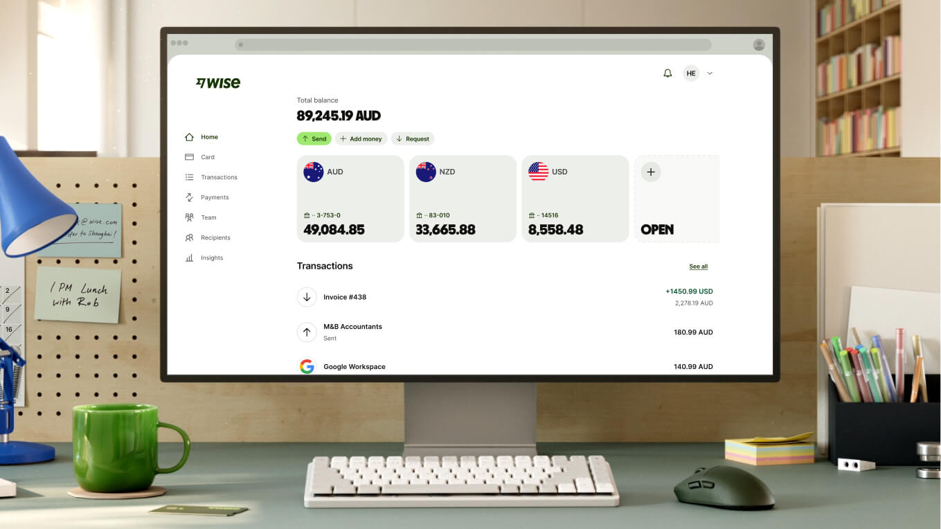 A close-up shot of a modern workspace shows a white computer monitor displaying the Wise dashboard. The screen shows a total balance of "89,245.19 AUD" and three main currency accounts: AUD with "49,084.85," NZD with "33,665.88," and USD with "8,558.48," followed by an option to open a new account. Recent transactions are listed below. In front of the monitor is a compact white keyboard, a dark grey mouse, and a bright green ceramic mug.